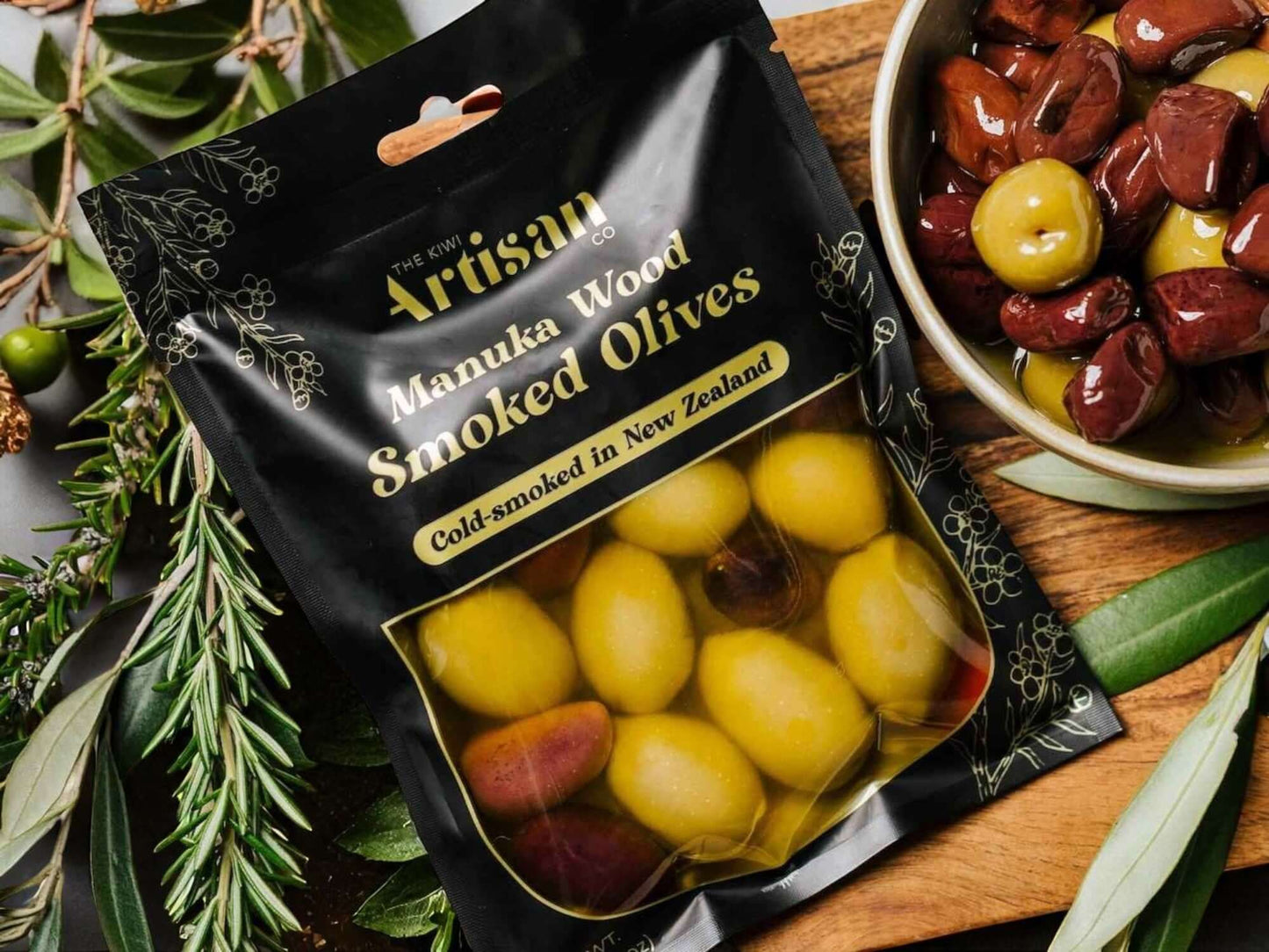 Packaged Manuka Wood Smoked Olives with a bowl of olives on a wooden surface.