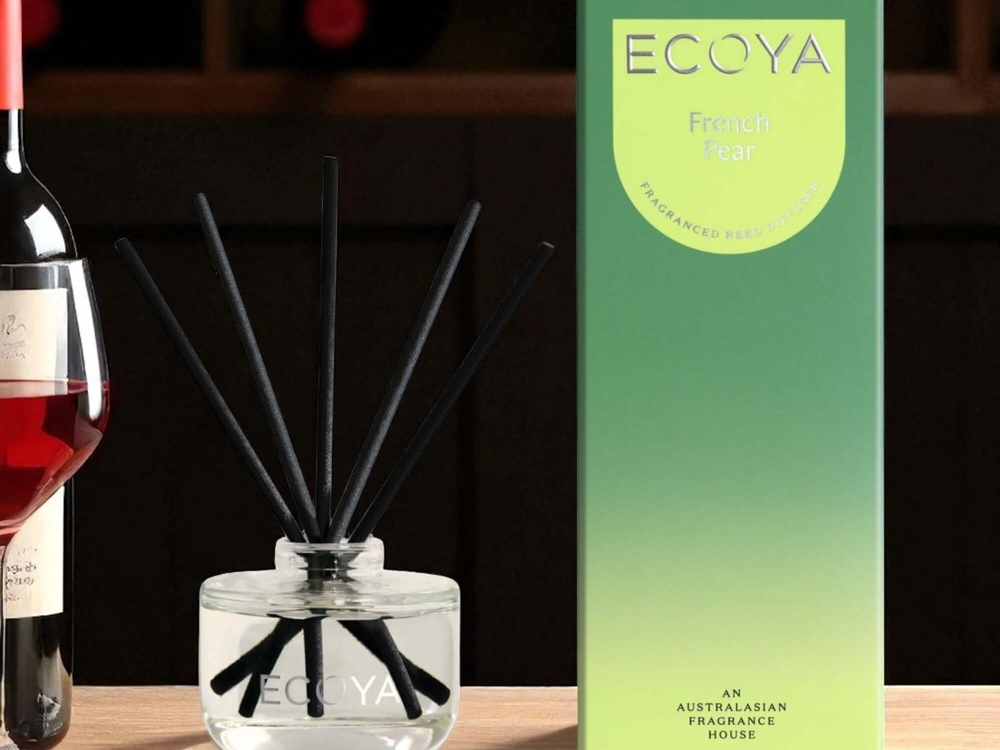 Ecoya diffuser with black sticks next to a bottle of wine and its packaging on a wooden surface.