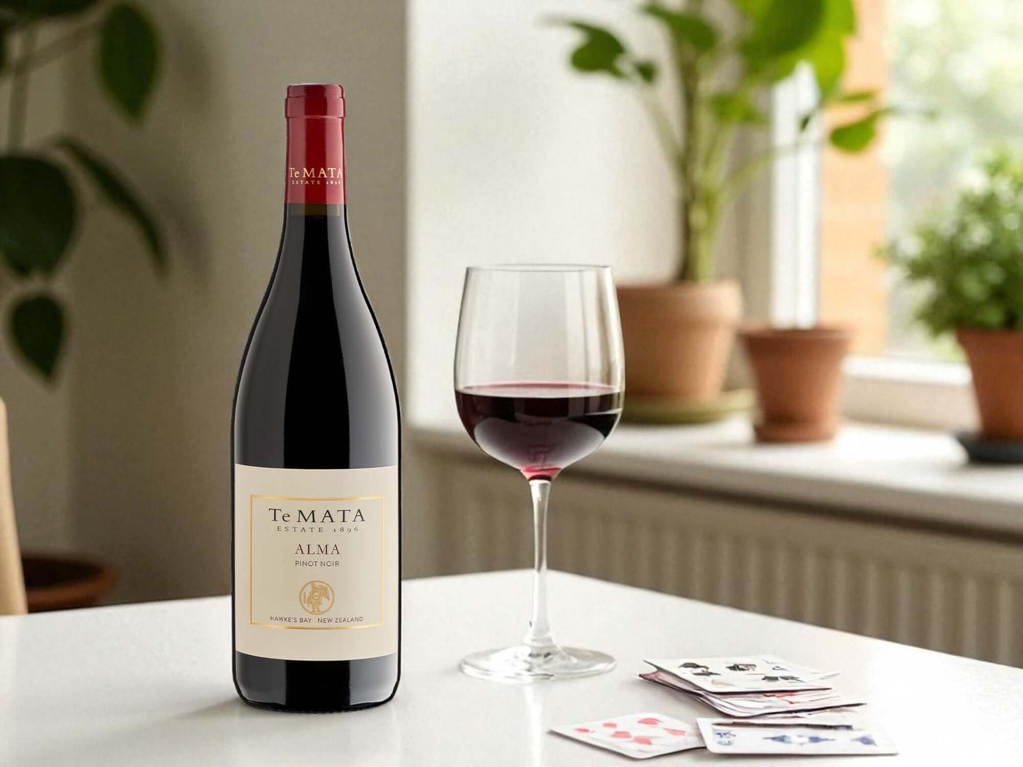 Te Mata Alma wine bottle with a glass of wine on a table with playing cards, plants in the background