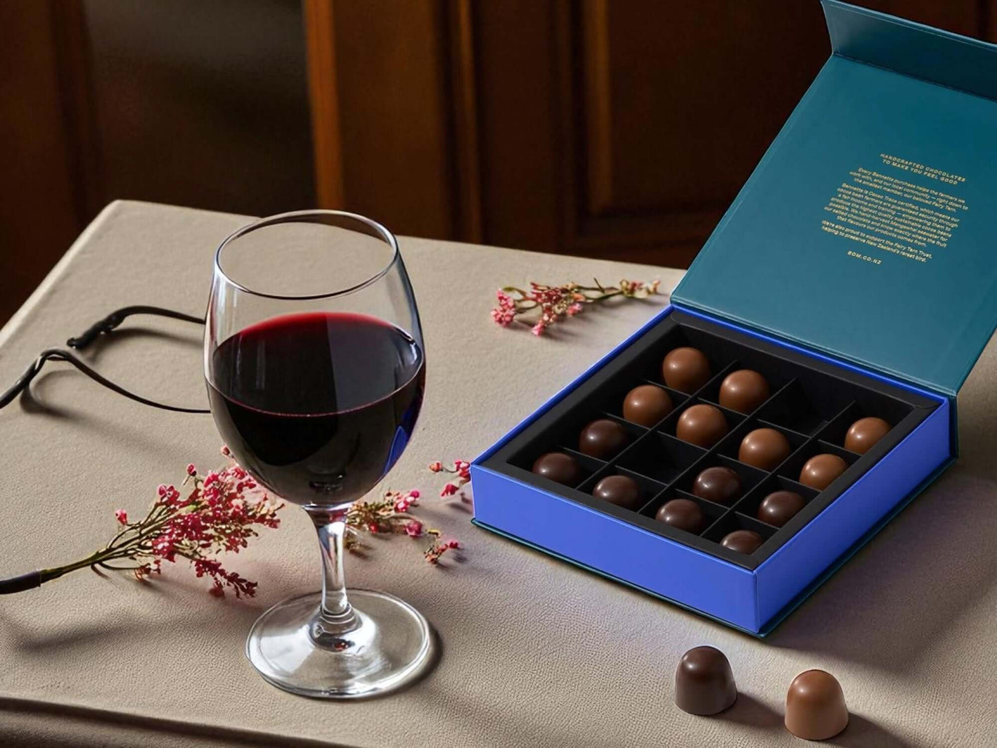 Glass of red wine next to a box of chocolates on a table with pink flowers.