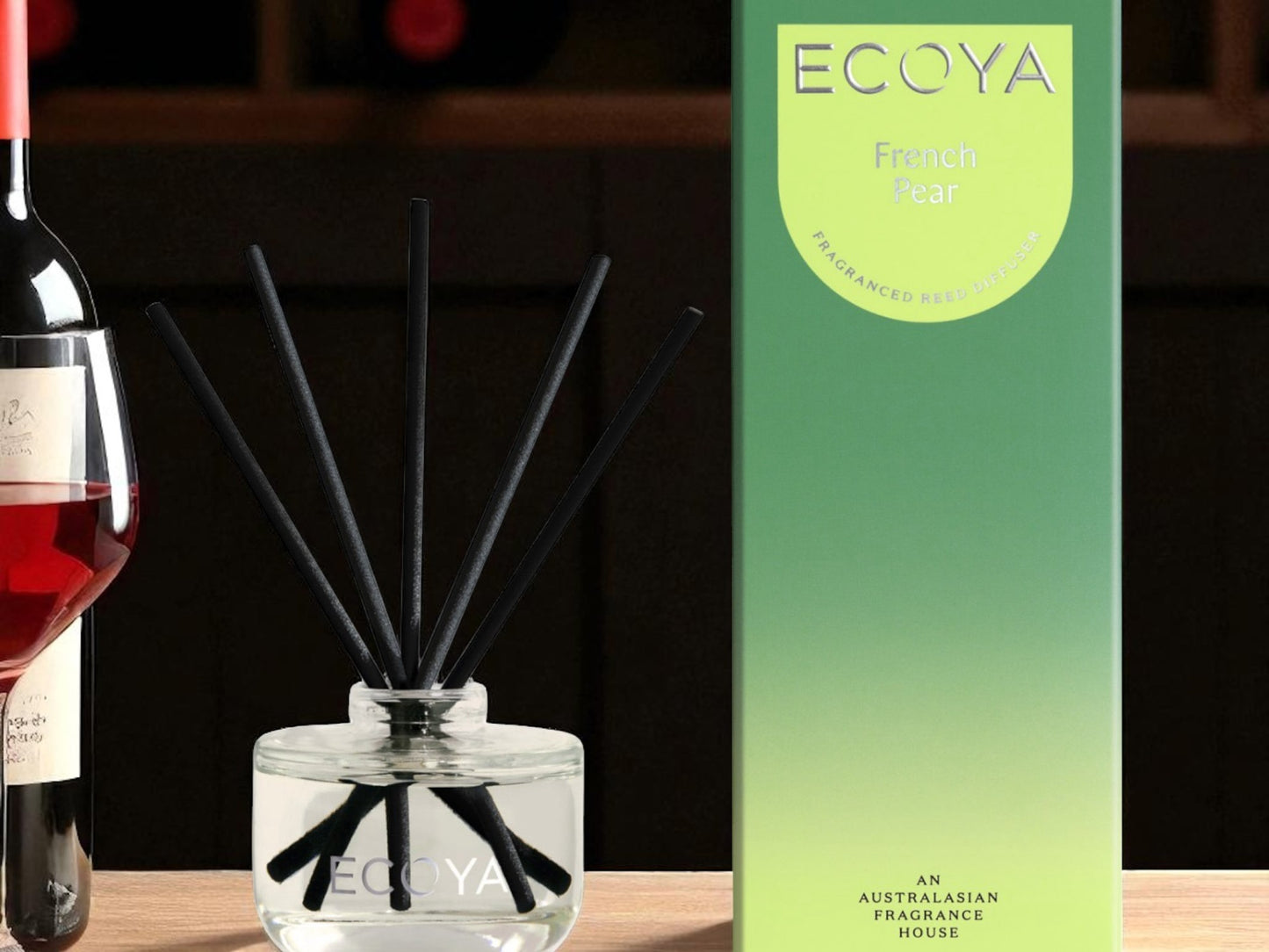 Ecoya diffuser with black sticks next to a bottle of wine and its packaging on a wooden surface.