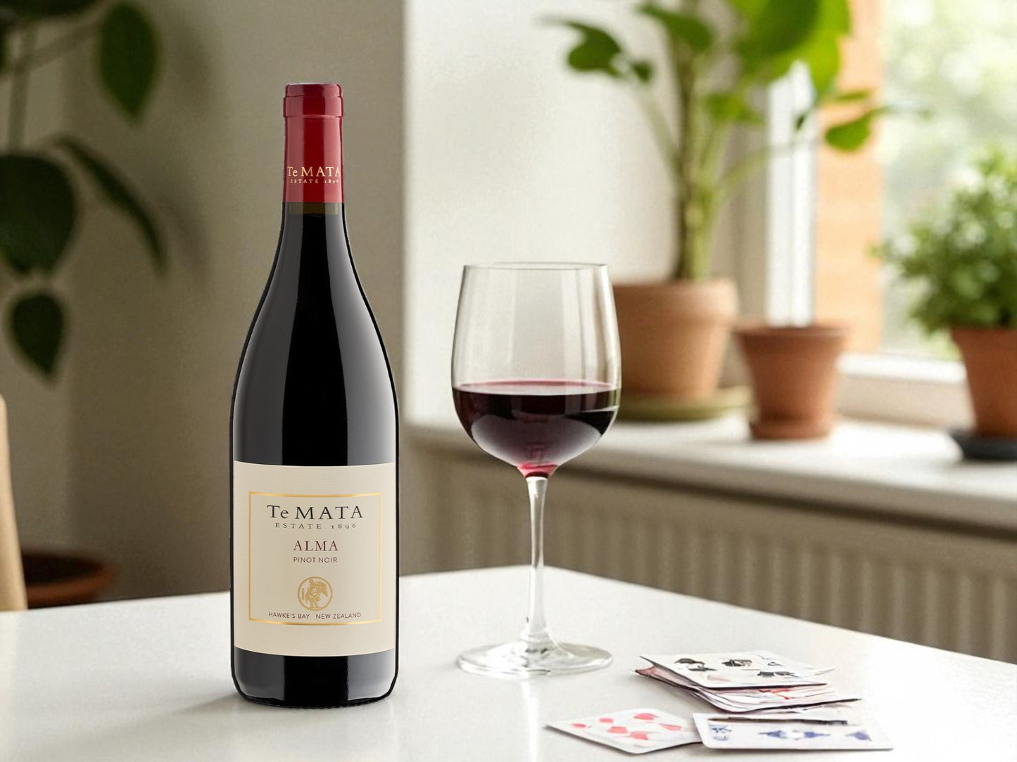 Te Mata Alma wine bottle with a glass of wine on a table with playing cards, plants in the background
