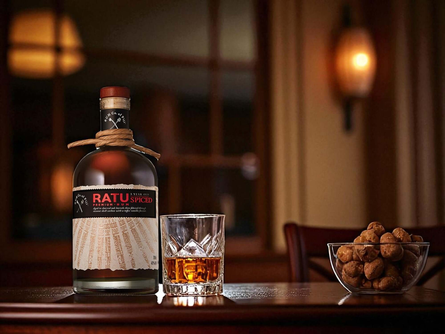 Bottle of Ratu rum with a glass and bowl of nuts on a wooden table.
